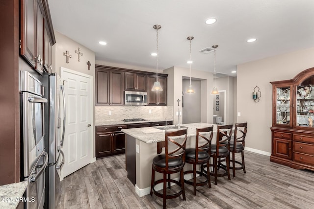 kitchen with pendant lighting, wood-type flooring, a kitchen island with sink, stainless steel appliances, and light stone countertops