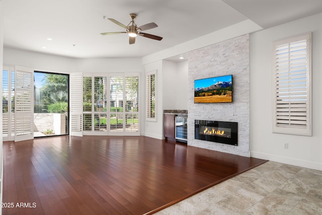 unfurnished living room featuring ceiling fan, a stone fireplace, wine cooler, wood finished floors, and baseboards