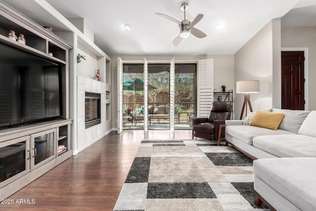 living room with a tiled fireplace, ceiling fan, dark hardwood / wood-style flooring, and built in shelves