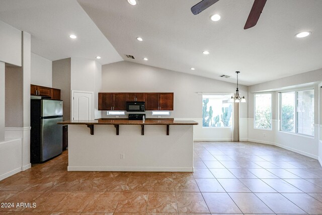 kitchen with light tile patterned floors, ceiling fan with notable chandelier, stainless steel refrigerator, a kitchen bar, and a kitchen island with sink