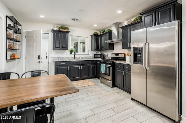 kitchen with tasteful backsplash, appliances with stainless steel finishes, light stone counters, and wall chimney exhaust hood