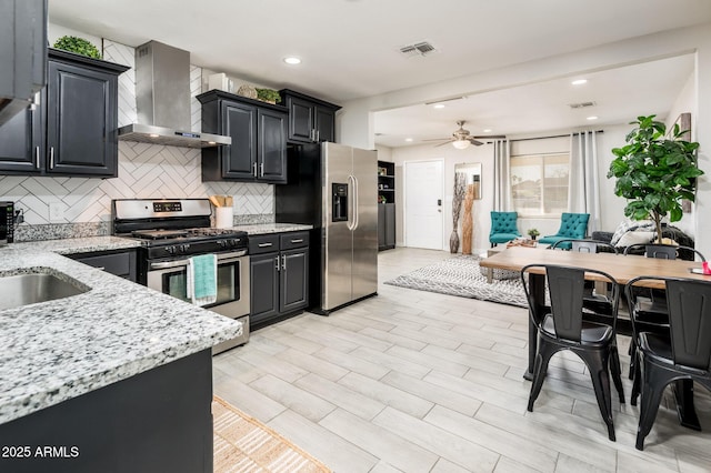 kitchen with wall chimney exhaust hood, light stone counters, ceiling fan, stainless steel appliances, and decorative backsplash