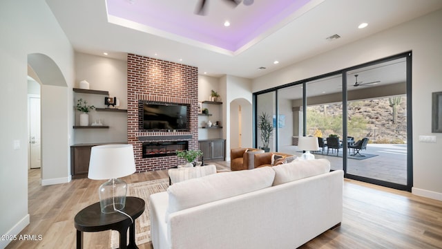 living room with a brick fireplace, a tray ceiling, light hardwood / wood-style floors, and ceiling fan