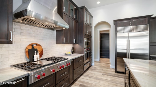 kitchen featuring dark brown cabinetry, ventilation hood, appliances with stainless steel finishes, light hardwood / wood-style floors, and backsplash