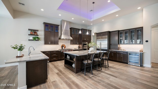 kitchen with a breakfast bar area, a tray ceiling, kitchen peninsula, beverage cooler, and wall chimney range hood