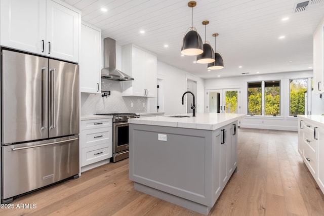 kitchen with sink, appliances with stainless steel finishes, a kitchen island with sink, white cabinetry, and wall chimney exhaust hood