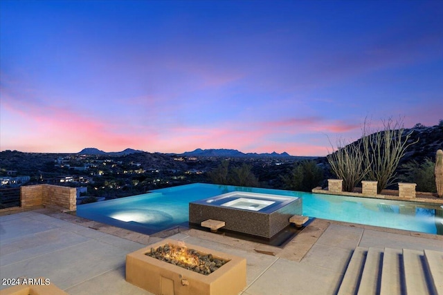 pool at dusk with an in ground hot tub, a mountain view, an outdoor fire pit, and a patio area