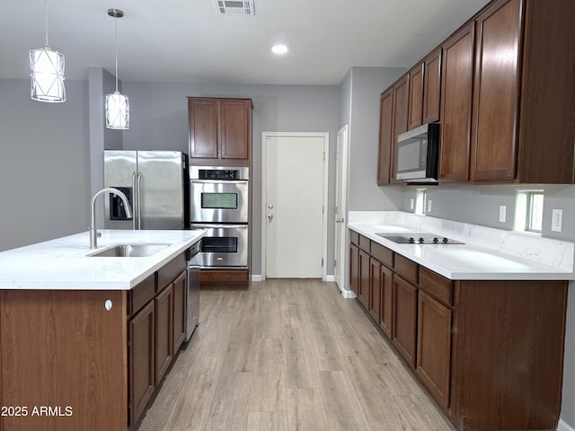 kitchen with light wood finished floors, a sink, stainless steel appliances, a center island with sink, and light countertops