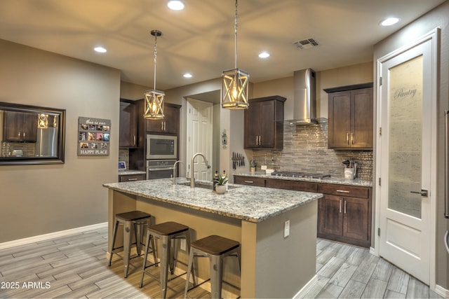 kitchen featuring light stone counters, wall chimney exhaust hood, a kitchen island with sink, appliances with stainless steel finishes, and sink