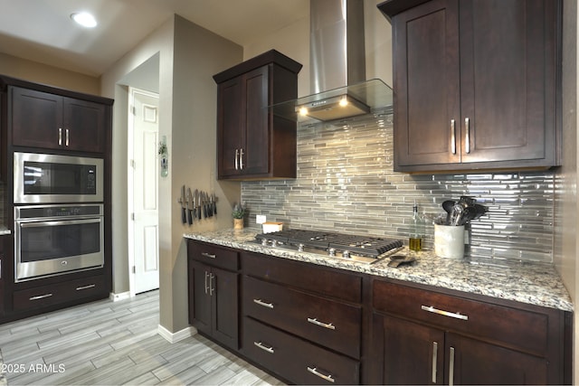 kitchen featuring stainless steel appliances, light stone counters, decorative backsplash, dark brown cabinetry, and wall chimney range hood