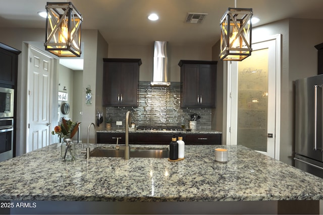 kitchen featuring stainless steel appliances, an island with sink, wall chimney range hood, tasteful backsplash, and dark brown cabinetry