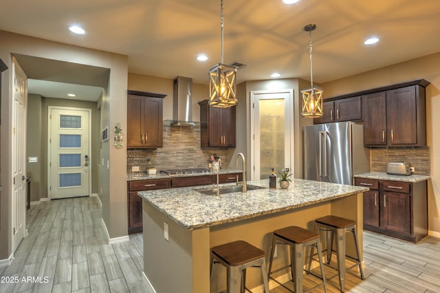 kitchen featuring stainless steel refrigerator, a kitchen island with sink, wall chimney range hood, and backsplash