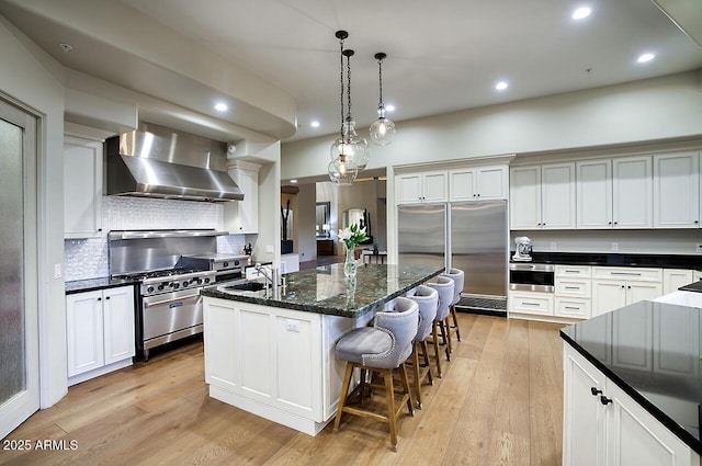 kitchen with wall chimney exhaust hood, a kitchen island with sink, stainless steel appliances, light wood-style floors, and a sink