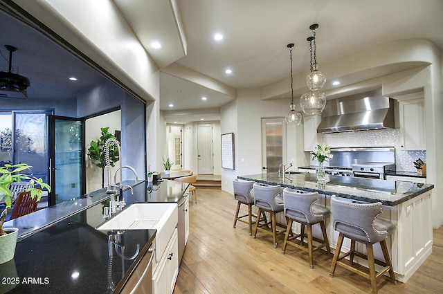 kitchen with light wood-type flooring, wall chimney exhaust hood, dark stone counters, and a sink