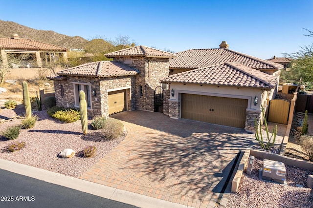 mediterranean / spanish house featuring stone siding, a tiled roof, an attached garage, a gate, and decorative driveway