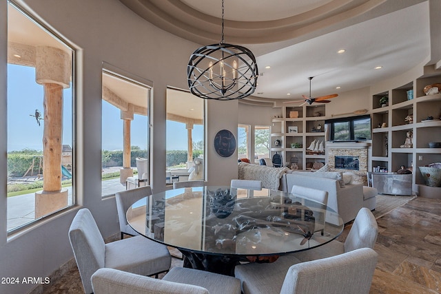 dining room with ceiling fan with notable chandelier, a stone fireplace, a tray ceiling, and built in features