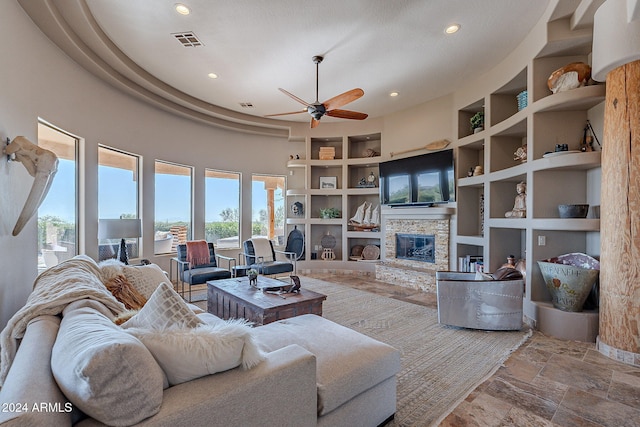 living room featuring ceiling fan, a textured ceiling, a high ceiling, built in shelves, and a stone fireplace