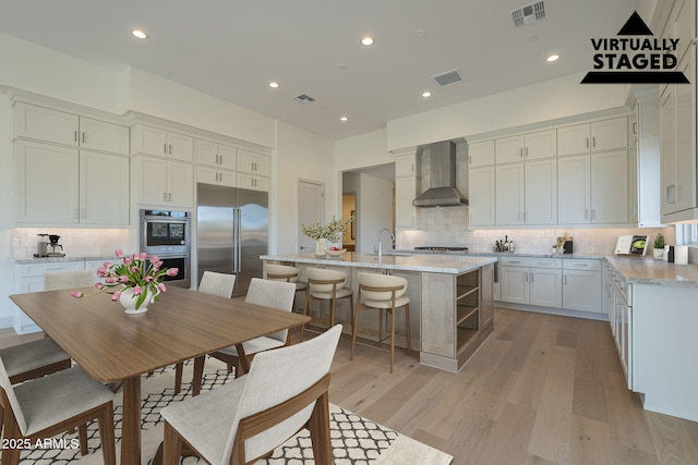 kitchen featuring sink, appliances with stainless steel finishes, an island with sink, light hardwood / wood-style floors, and wall chimney range hood