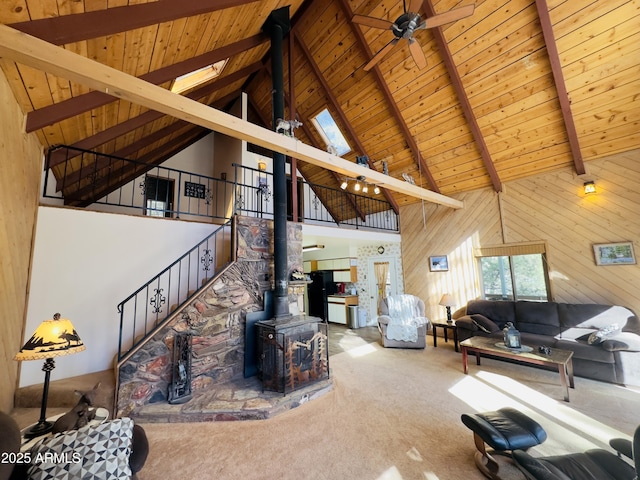 living room featuring wood walls, a wood stove, carpet flooring, beamed ceiling, and ceiling fan