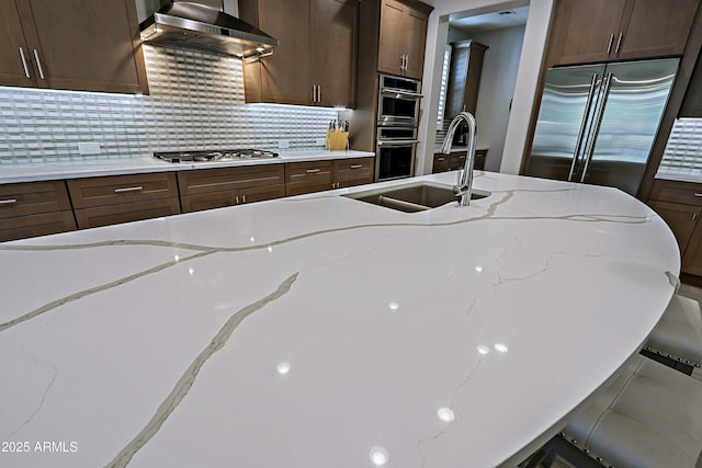 kitchen featuring wall chimney exhaust hood, sink, tasteful backsplash, stainless steel appliances, and light stone countertops