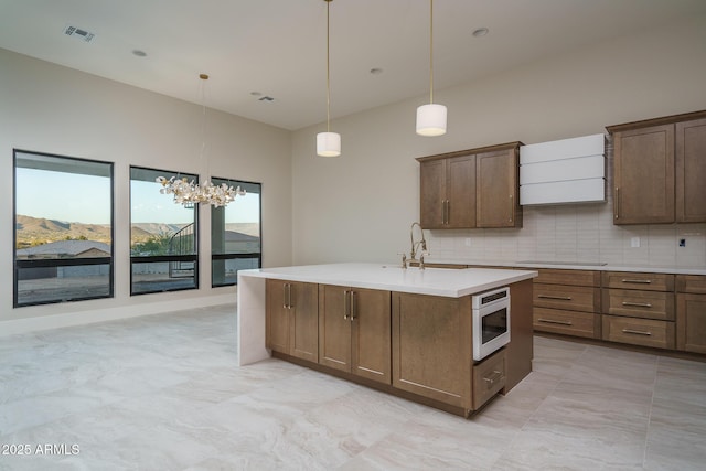 kitchen with tasteful backsplash, hanging light fixtures, stainless steel microwave, a mountain view, and a kitchen island with sink