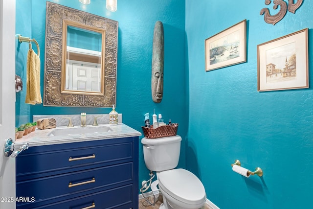 half bathroom featuring baseboards, a textured wall, vanity, and toilet