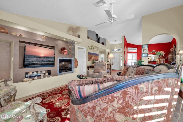 living room with ceiling fan, a stone fireplace, a textured ceiling, and high vaulted ceiling