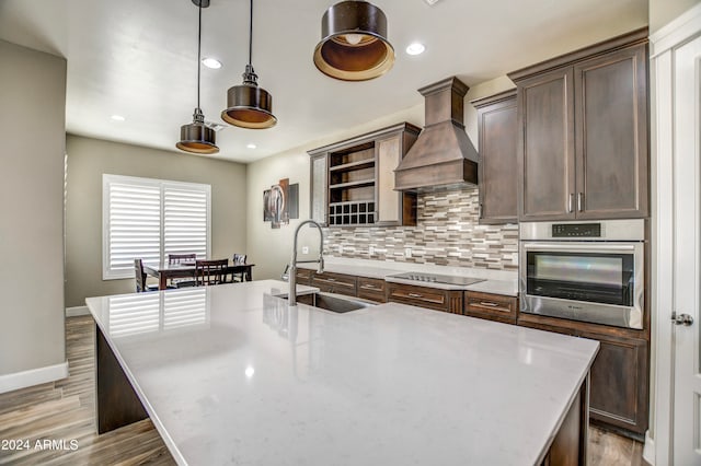 kitchen with black electric stovetop, stainless steel oven, custom range hood, pendant lighting, and sink