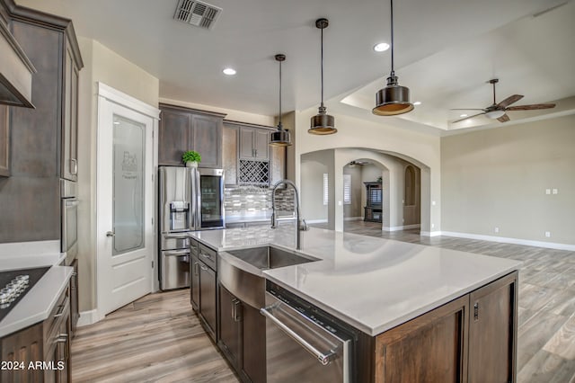 kitchen with dark brown cabinetry, stainless steel appliances, an island with sink, hanging light fixtures, and ceiling fan