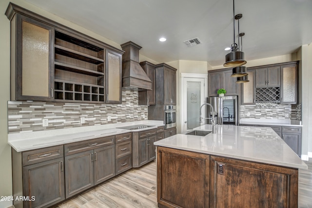 kitchen with dark brown cabinetry, hanging light fixtures, an island with sink, stainless steel appliances, and custom range hood