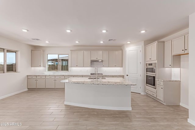 8553 N 175th Ln, Waddell, AZ 85355 - kitchen featuring light wood-style floors, stainless steel double oven, an island with sink, light stone countertops, and baseboards