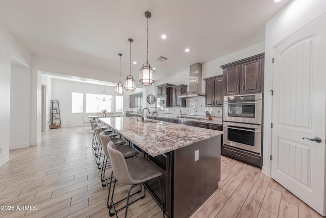 kitchen featuring stainless steel appliances, wall chimney range hood, a large island with sink, dark brown cabinets, and sink