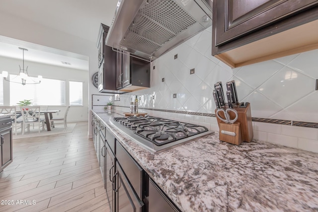 kitchen featuring decorative light fixtures, wall chimney range hood, an inviting chandelier, stainless steel gas cooktop, and dark brown cabinets