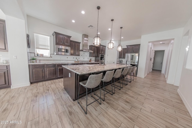 kitchen featuring hanging light fixtures, wall chimney range hood, a breakfast bar, a kitchen island with sink, and appliances with stainless steel finishes