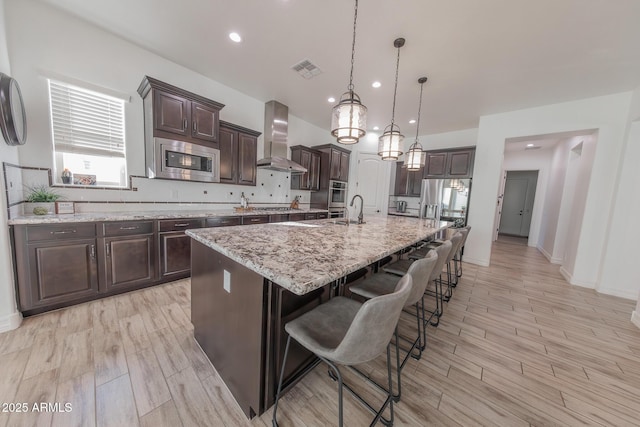 kitchen with a large island with sink, dark brown cabinets, hanging light fixtures, wall chimney range hood, and appliances with stainless steel finishes