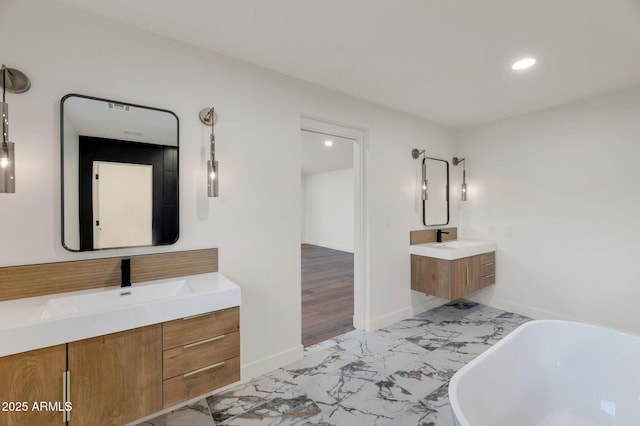 full bath featuring baseboards, a sink, marble finish floor, a freestanding bath, and two vanities