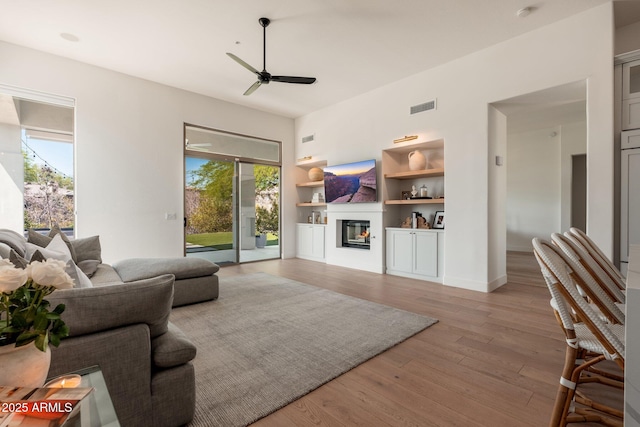 living room with built in shelves, a ceiling fan, a glass covered fireplace, light wood finished floors, and baseboards