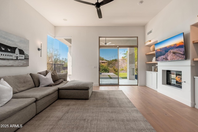 living area featuring a ceiling fan, a glass covered fireplace, wood finished floors, baseboards, and built in shelves