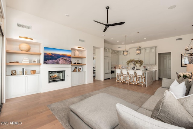 living area featuring ceiling fan, light wood-style flooring, a glass covered fireplace, built in shelves, and recessed lighting