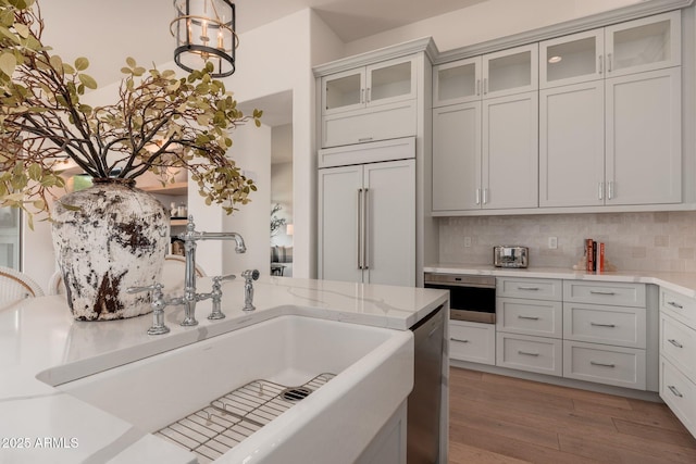 kitchen featuring a sink, paneled built in refrigerator, wood finished floors, backsplash, and glass insert cabinets