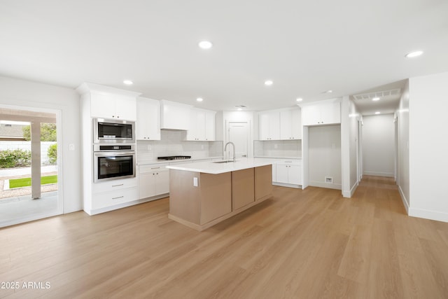 kitchen with appliances with stainless steel finishes, a sink, decorative backsplash, a center island with sink, and light wood-style flooring
