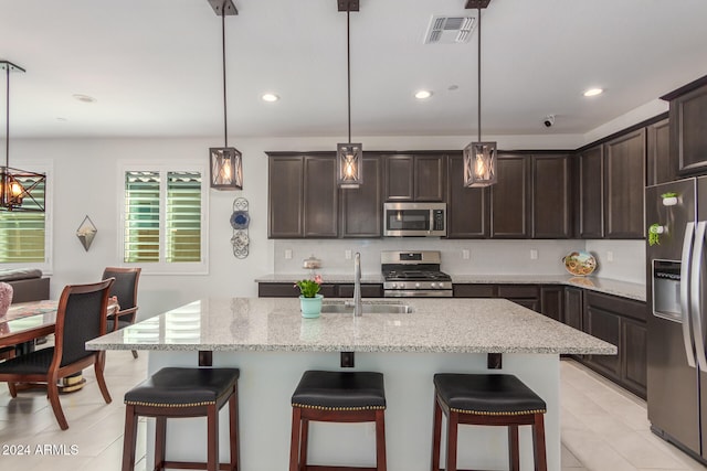 kitchen featuring a center island with sink, sink, light stone counters, appliances with stainless steel finishes, and pendant lighting