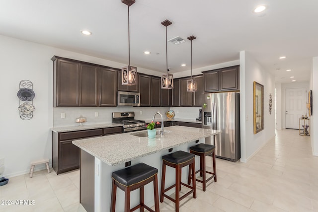 kitchen featuring a center island with sink, light stone counters, hanging light fixtures, dark brown cabinets, and appliances with stainless steel finishes