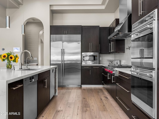 kitchen featuring light countertops, decorative backsplash, a sink, wall chimney range hood, and built in appliances