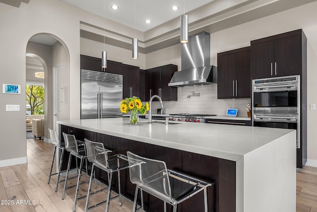 kitchen featuring an island with sink, a breakfast bar area, stainless steel appliances, light countertops, and wall chimney range hood