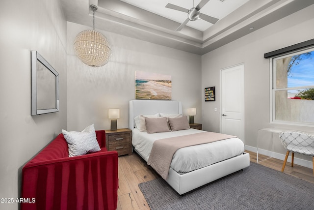 bedroom with ceiling fan with notable chandelier, a tray ceiling, wood finished floors, and baseboards