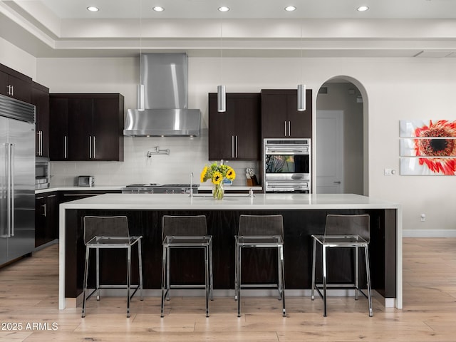 kitchen with wall chimney exhaust hood, a kitchen island with sink, and light countertops