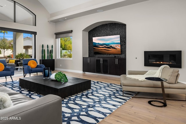 living area featuring light wood finished floors, baseboards, high vaulted ceiling, and a glass covered fireplace