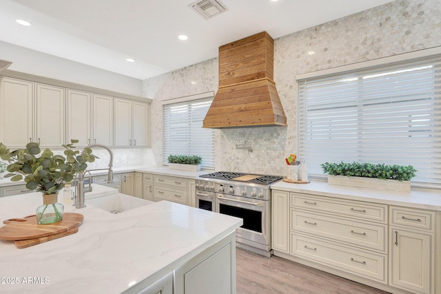 kitchen featuring sink, custom exhaust hood, light stone countertops, cream cabinets, and range with two ovens