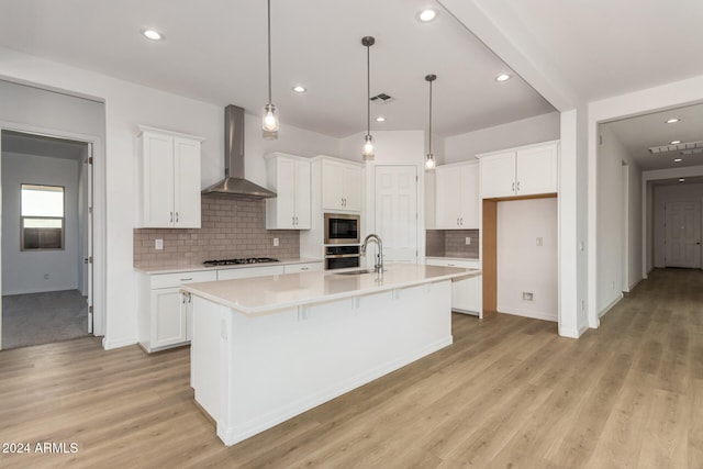 kitchen with wall chimney range hood, stainless steel appliances, white cabinets, light hardwood / wood-style flooring, and a kitchen island with sink